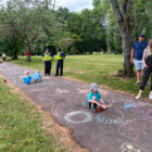 three children draw in chalk on path while parents and two police officer watch