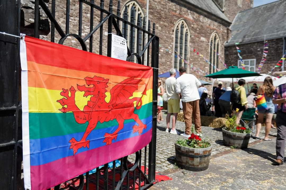 a pride rainbow flag with a welsh dragon on it- draped on a church gate