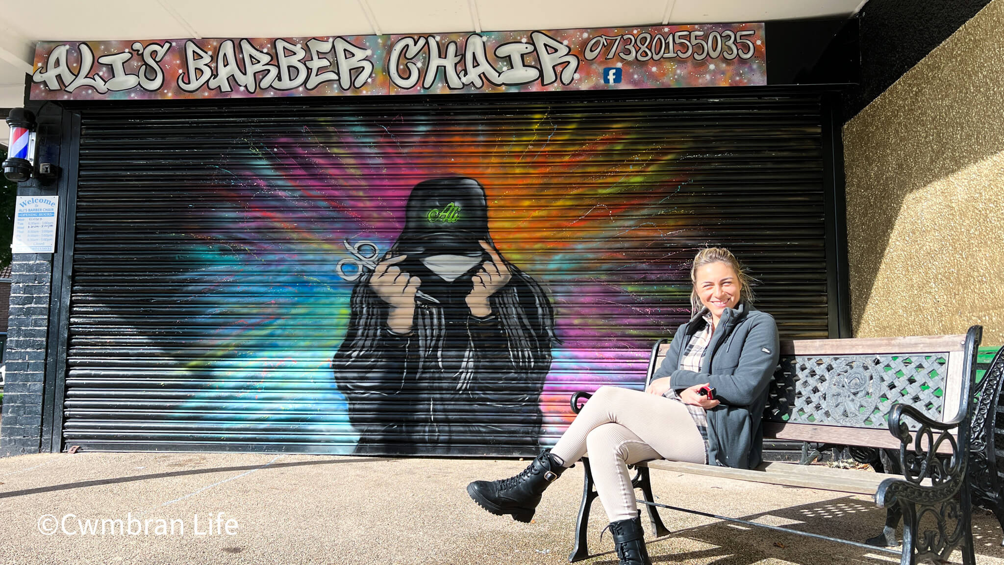 woman sat on bench outside a barbers- the shutters are covered in graffiti