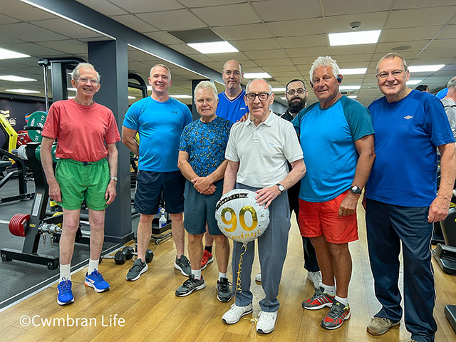 group of men in a gym- one is 90 and holds a birthday balloon