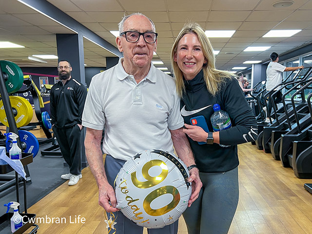 man in gym with woman- he's holding a 90th birthday balloon