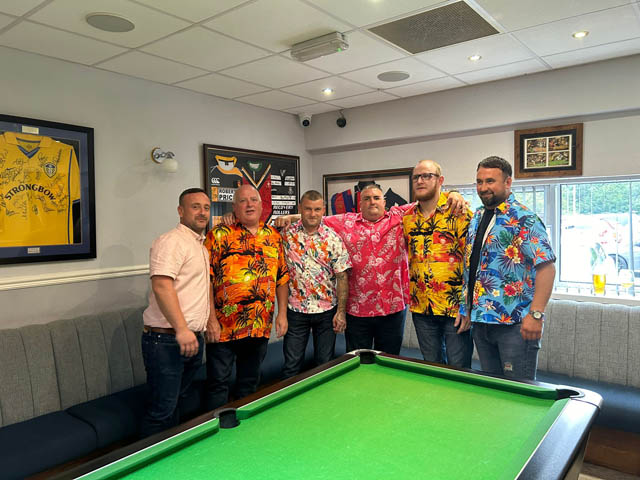 six men wearing Hawaiian shirts stood by a pool table