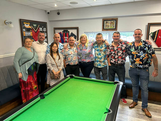 group of men and women, most wearing Hawaiian shirts , stood by a pool table