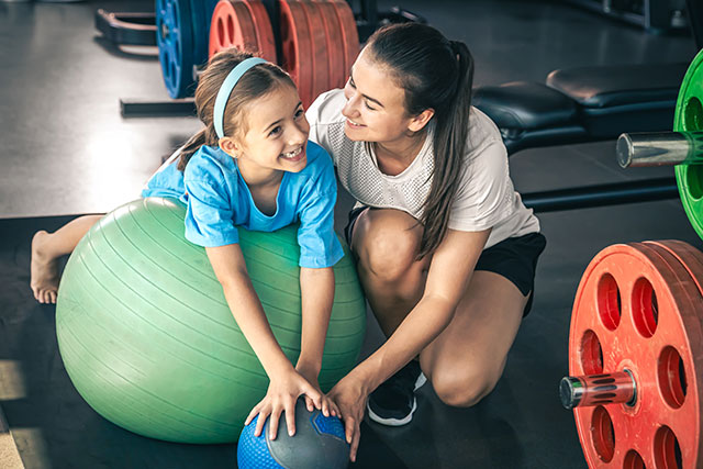 child girl stretching on pilates fitness ball with mum in gym.