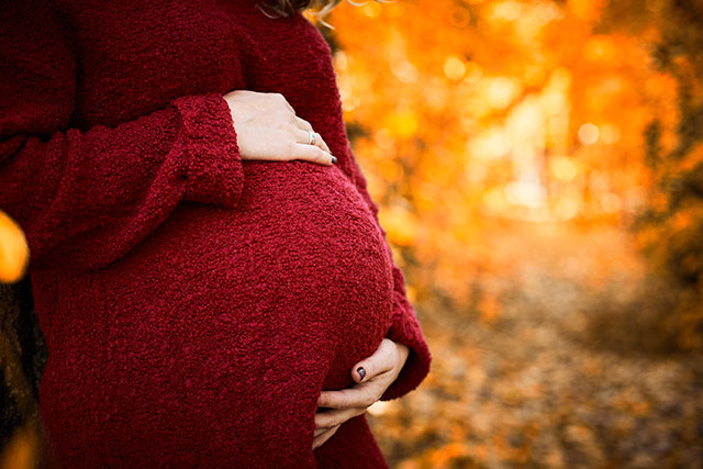 Unrecognizable young pregnant woman in red dress standing in forest among autumn leaves