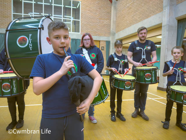 a boy blows in a shanter pipe wth four drummers behind him