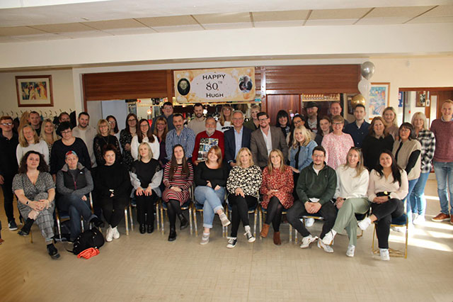 a large group of staff smile in front of an 80th birthday sign