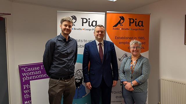 two men and a woman in front of a banner saying Pia- the braille company