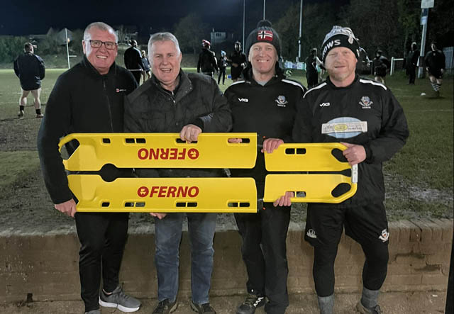 four men hold a stretcher on a rugby pitch