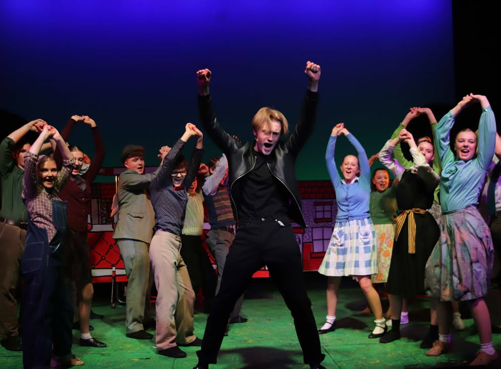 a group of young people in 1960s rock n roll clothes dance by a man in black leather trousers and jeans