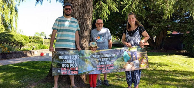 three adults and a young child hold a banner about dog poo in a park