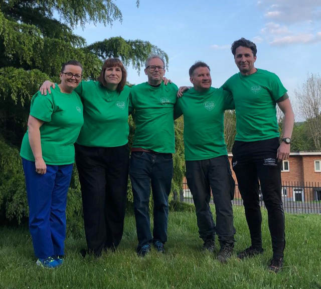 five adults in a row wearing green t-shirts smile at the camera