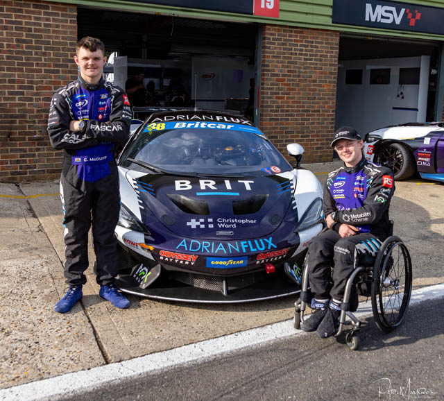 two teenage boys, one stood up and one in a wheelchair next to a racing car