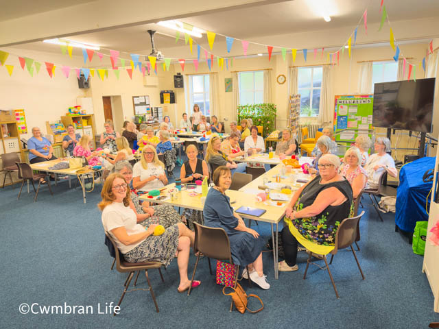 a roomful of women who are crocheting at tables smile for a photo