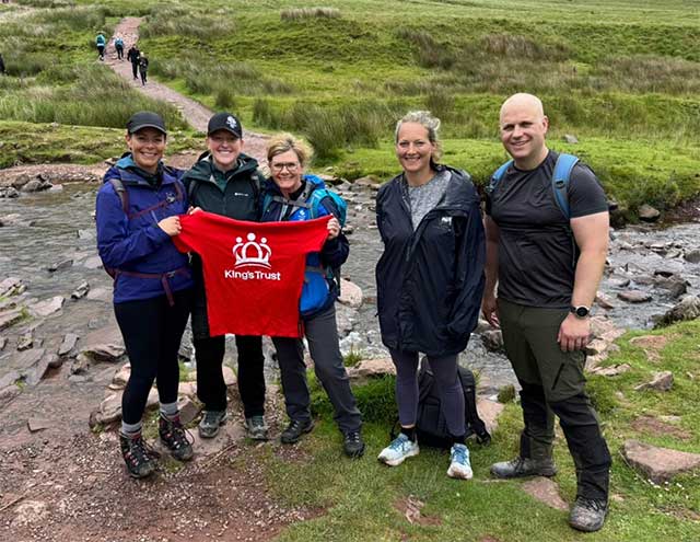 five people at the bottom of a mountain smile for a photo holding a red t shirt with the words The King's Trust on it