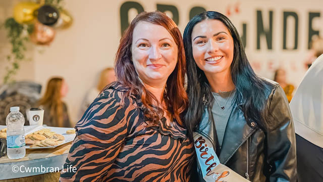 two women smile at the camera stood in cafe
