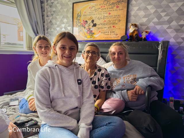 three adults women sit with their mum on a bed