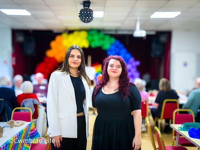 two women in a room in front of a balloon arch in colours of the rainbow