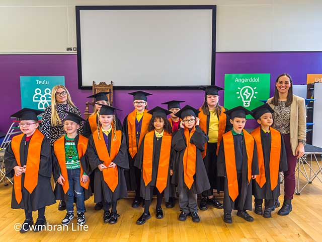 11 children wear graduation hat and gowns standing with two teachers