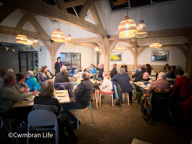 a room full of people sat at tables in cafe- one woman is stood up talking to them