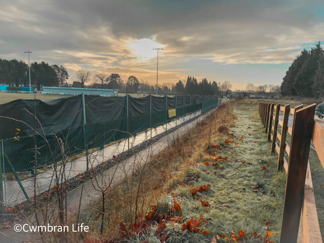 a fence near a football pitch- a black banner blocks the view for fans stood by the fence