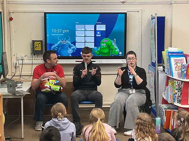 three adults sat in front of children- one women is doing sign language