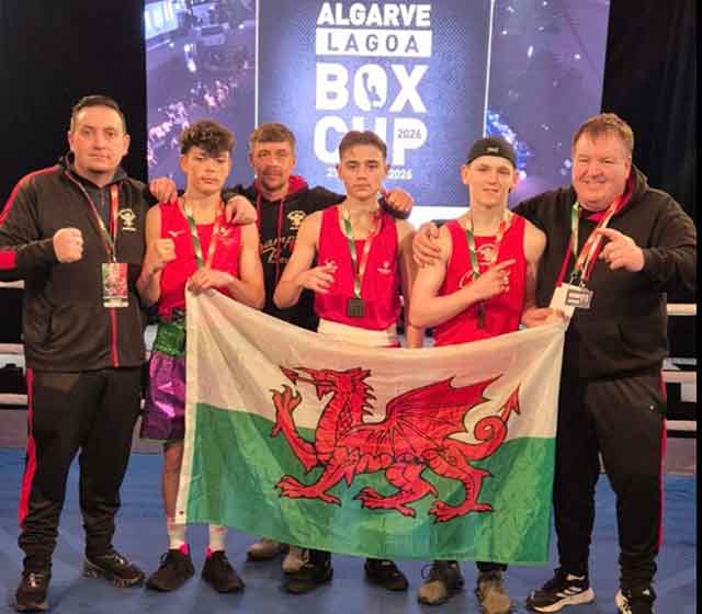 three teenage boys and three men in a boxing ring holding a wales flag