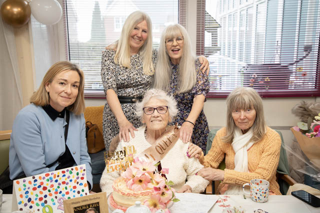 four women surround a woman sat at a table wearing a 100th birthday banner