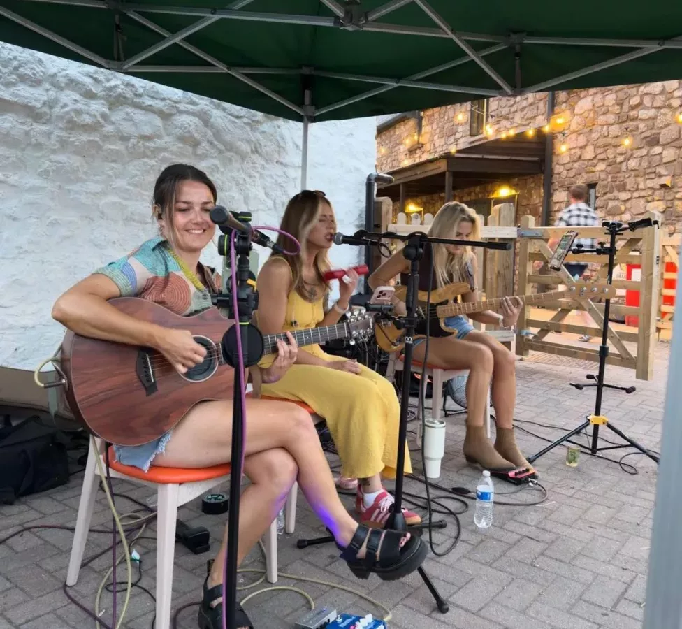 three women sat on chairs play guitars and sing